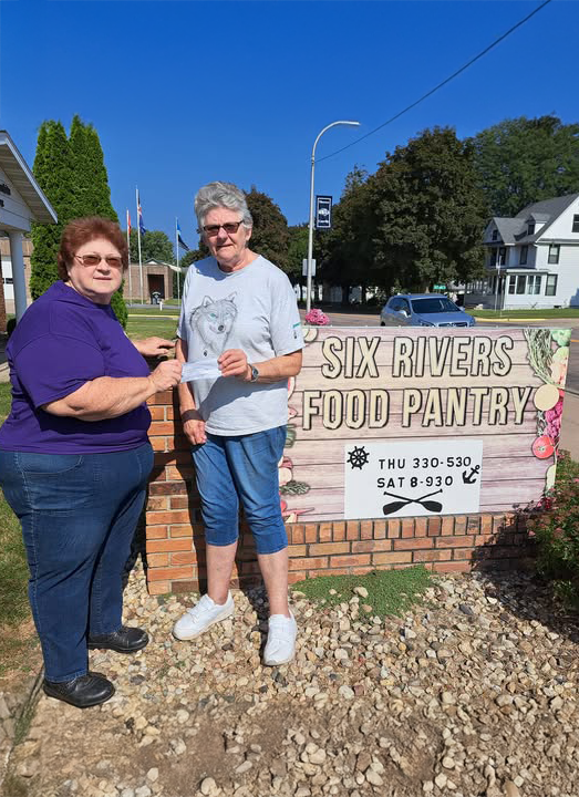 Two happy women holding a check in front of a sign that reads Six Rivers Food Pantry outside.