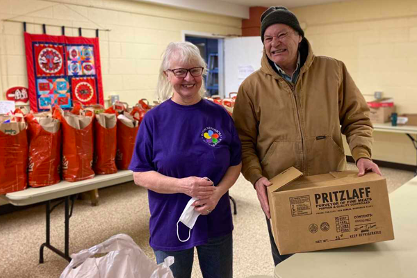 Photo of happy woman in purple t-shirt and happy man in tan coat holding a box in front of bagged items indoors.