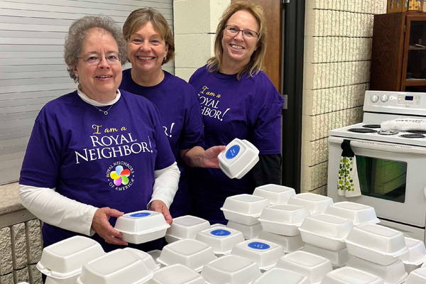 photo of Photo of three happy women wearing purple I am a Royal Neighbor shirts in a kitchen with styrofoam boxes.