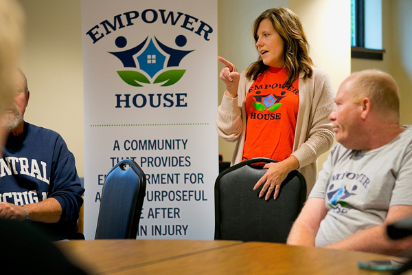 Woman in orange shirt speaking to people at a table indoors, looking at her, in front of an Empower House banner sign and descriptive text below.