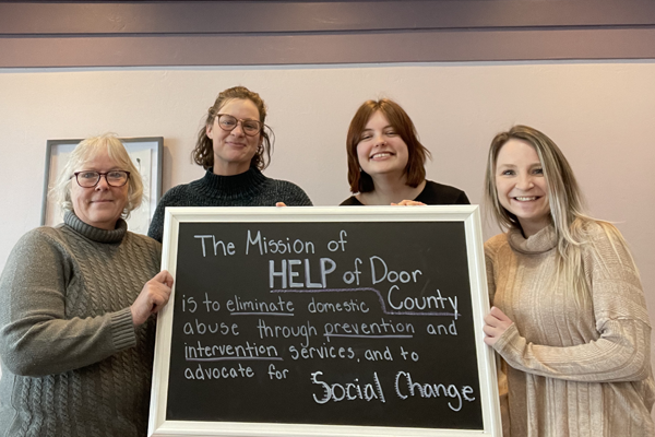 Four smiling women holding a chalkboard sign in a white frame. The chalkboard reads: The Mission of HELP of Door County is to eliminate domestic abuse through prevention and intervention services, and to advocate for Social Change.