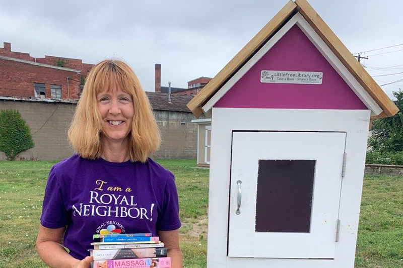 Smiling woman in I am a Royal Neighbors purple t-shirt holding books in front of a Little Free Library box outside.