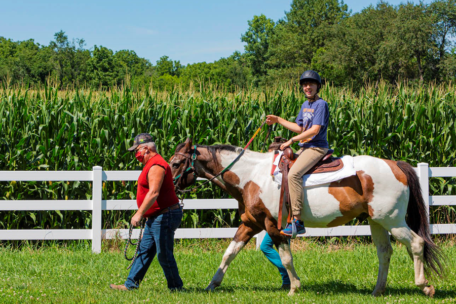 Photo of man leading a white and brown horse with a child student on the back by a white fence and cornfield.