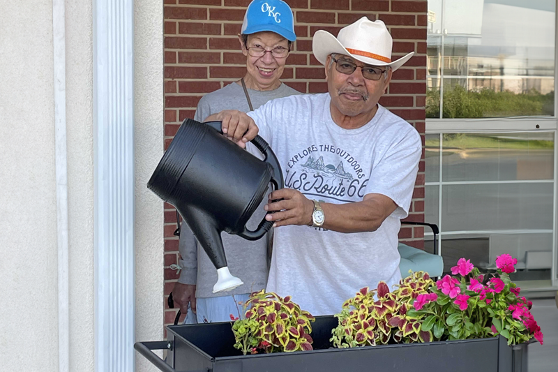 Smiling woman standing behind a man holding a watering can over plants outside in front of a brick building.