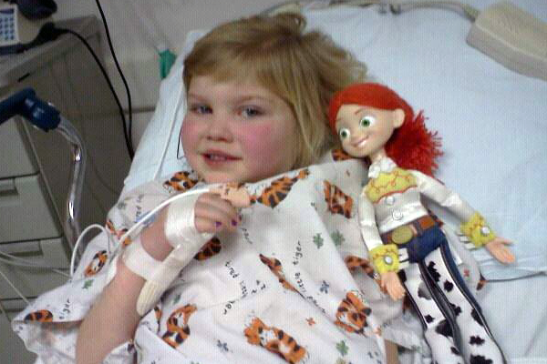 Smiling blonde girl in hospital gown with a cowgirl doll laying in a hospital bed.