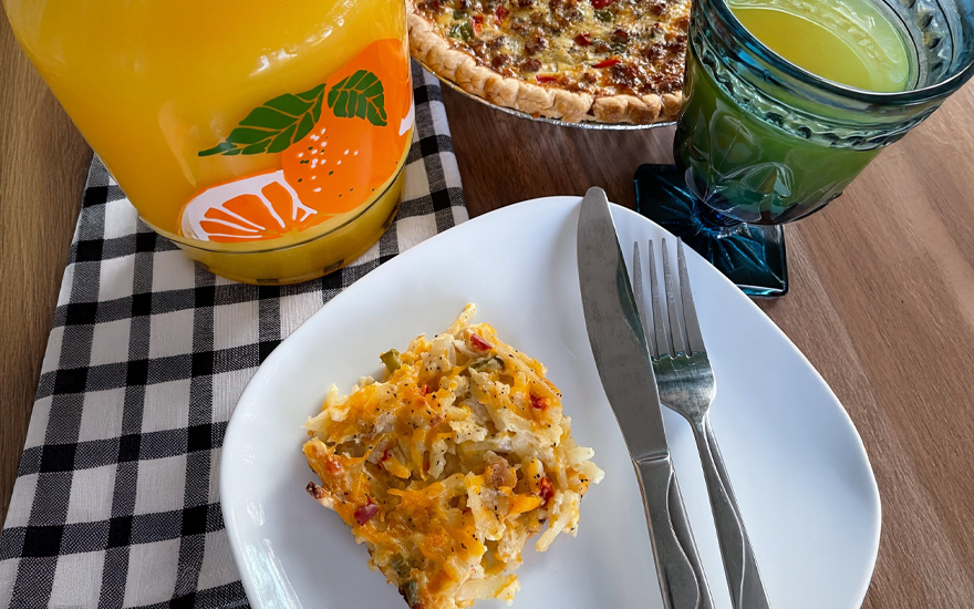 Orange juice bottle and in a blue glass next to a breakfast casserole with a piece of the casserole on a plate on a wooden tale.