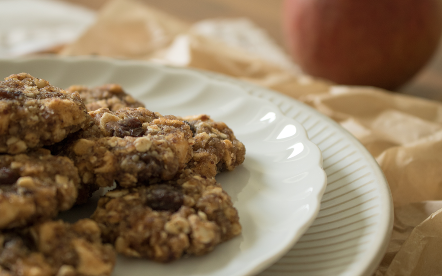 Close-up of Apple Raisin Cookies on a white plate in foreground and a red apple in background on a table.