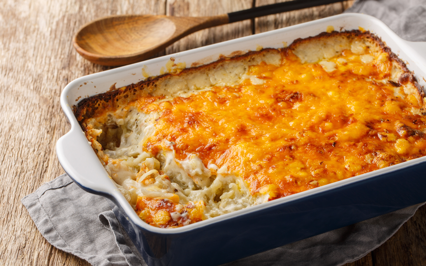 Close up of cheesy hash browns with a casserole dish with a wooden spoon nearby on a wooden table.