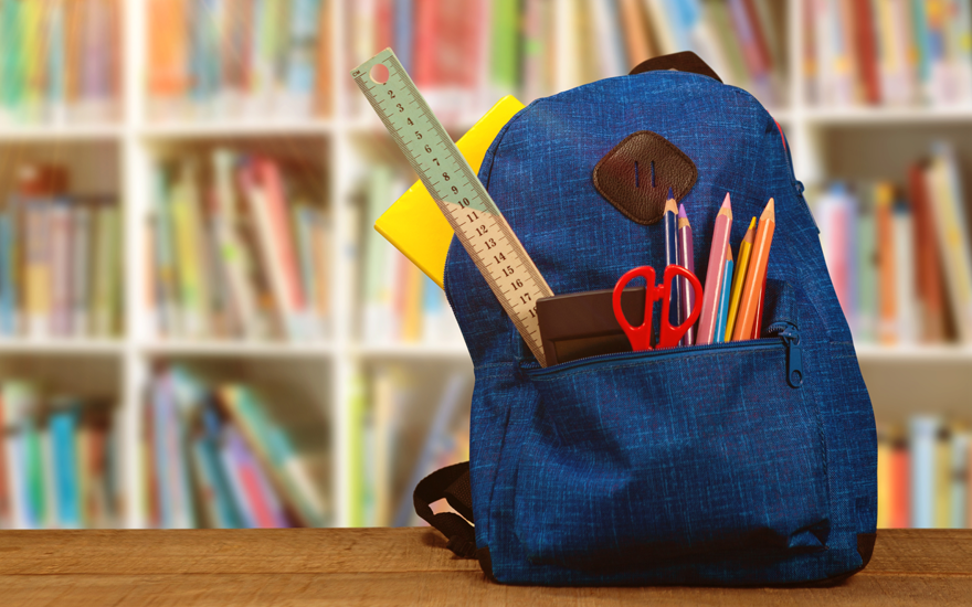 Blue backpack with school supplies on a table with blurred bookshelves with books background.