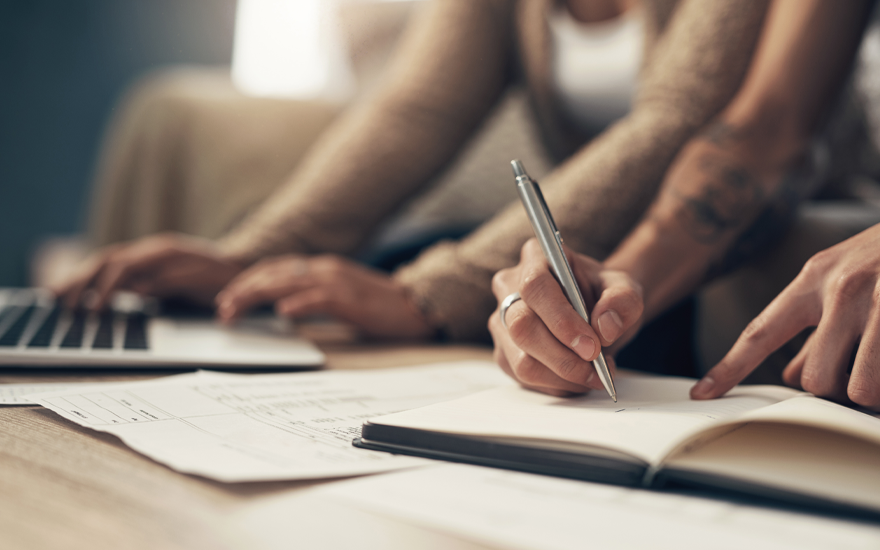 Close up writing in a notebook with paper and a laptop on a desk, two people blurred on a couch.
