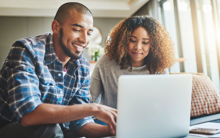 A happy couple sitting on a couch looking at a laptop.