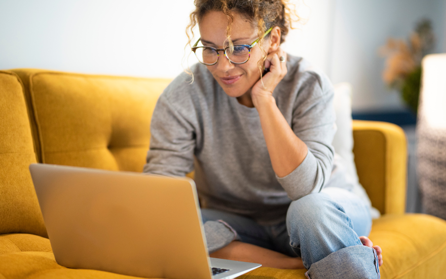 Smiling woman with curly hair pulled back and glasses, sitting on a yellow couch and looking at a laptop sitting on the couch.