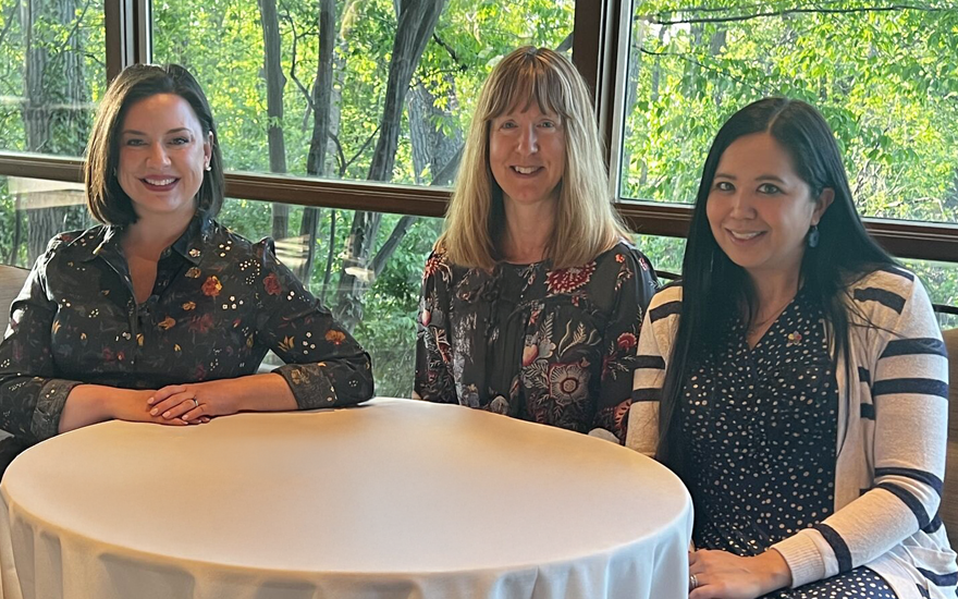 Three happy woman at a table with green woods behind outside of a large window.