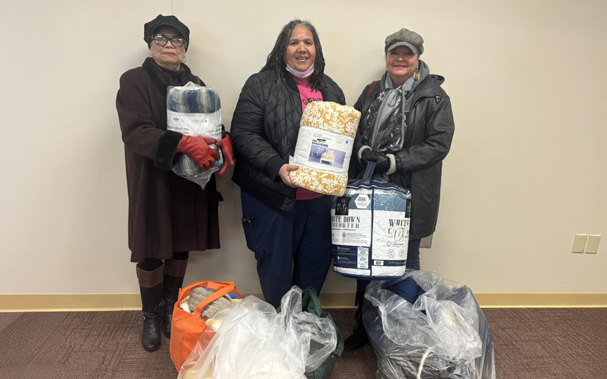 Three happy women in winter coats holding blankets with bags of blankets in front of them on the floor.