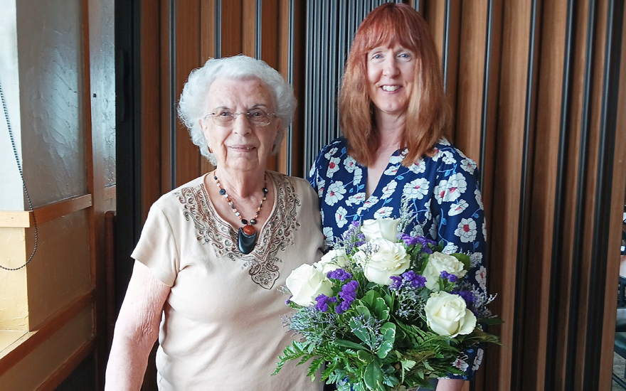 Two smiling women with a vase of white roses and purple flowers in front of them.