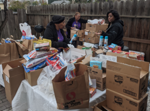 Three women packing boxes of food and toys outside on a table by a wooden fence.