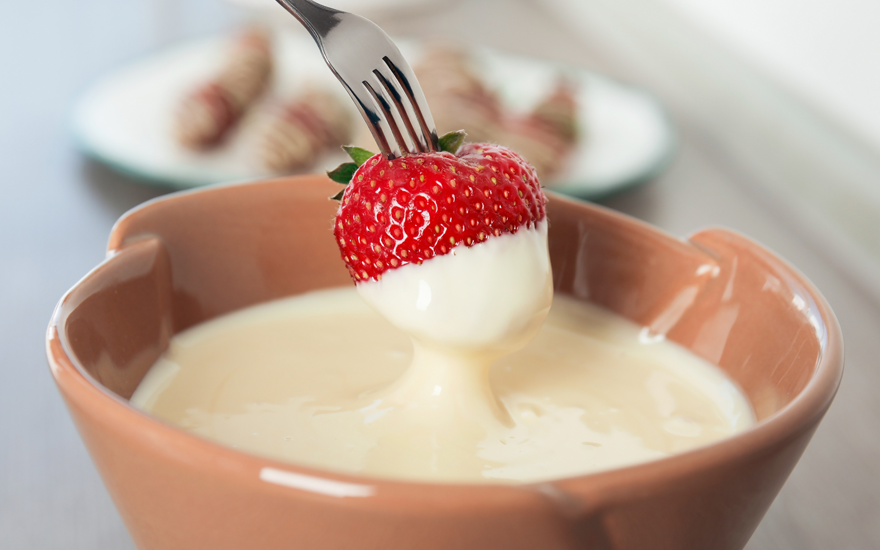 Close up of a strawberry on a fork being dipped into creamy dip in brown bowl.