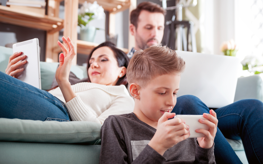 A dad on a laptop and a mom on a tablet on a couch with their son on a cell phone sitting on the floor in front of them.