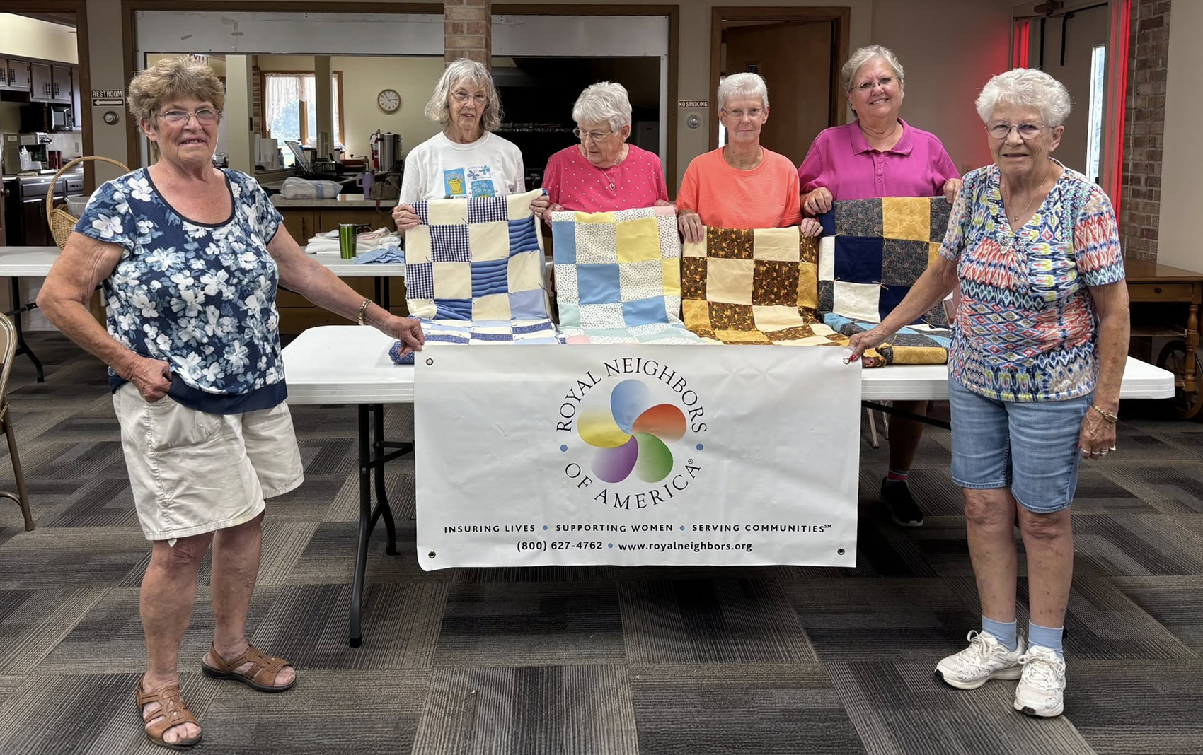 Six smiling women with quilts on a table that has a Royal Neighbors banner.