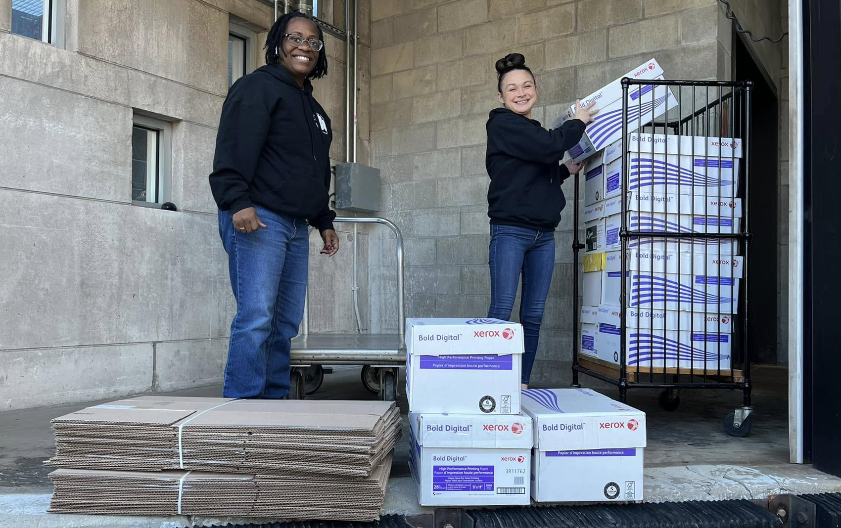 Two happy women loading cardboard boxes on a cart.