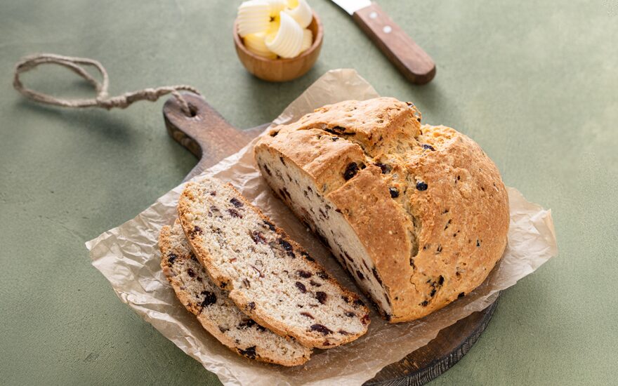 Sliced loaf of Irish soda bread on a cutting board next to a small bowl of butter and butter knife on a green table.