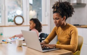 Mother on a laptop with daughter coloring in a kitchen.
