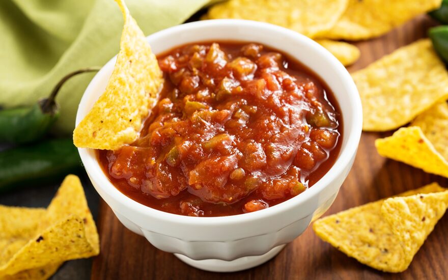 Close up of salsa in a small white bowl with a tortilla chip in it as well as scattered around the bowl.