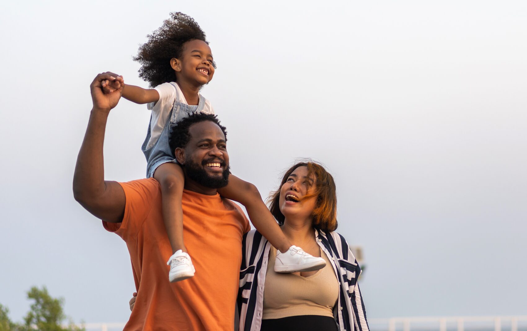 Happy family with young child with arm out on dad's shoulders next to mom outside.