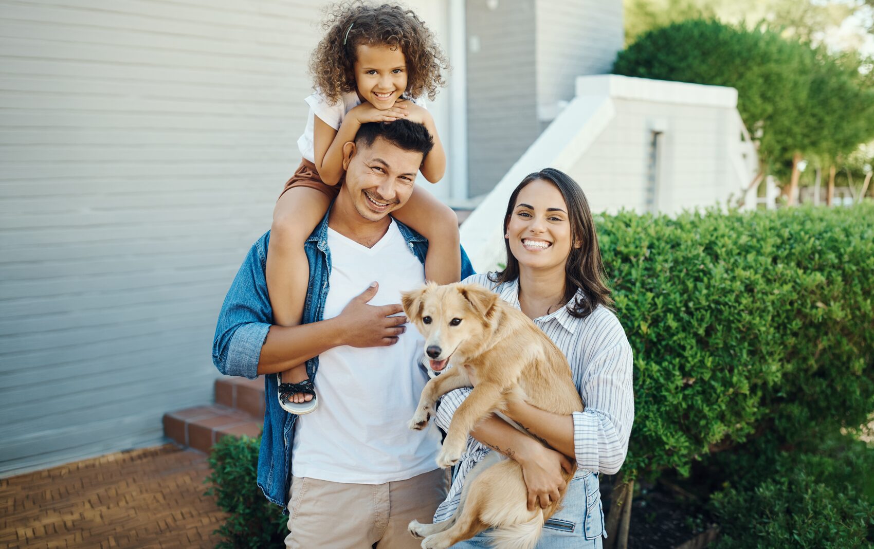 Happy family with a young daughter on dad's shoulders and mom holding a yellow dog outside.