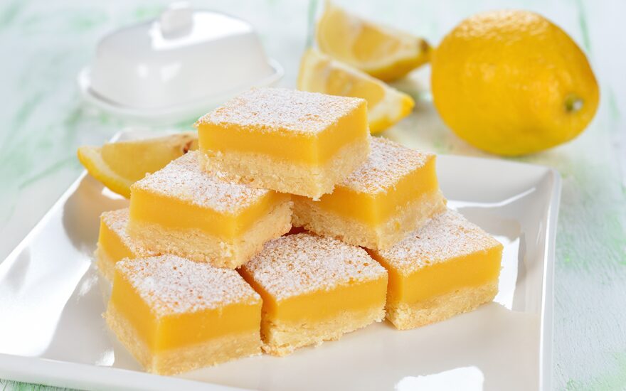 Plate of lemon bars stacked with sliced lemons in the background on a white plate.