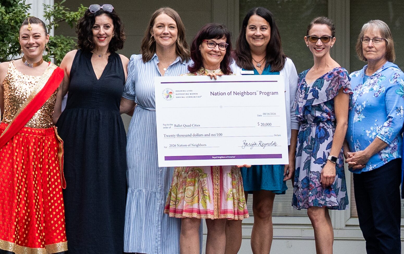 Group of happy women outside with the centered woman holding a big Nation of Neighbors $20.000 check on a stage.