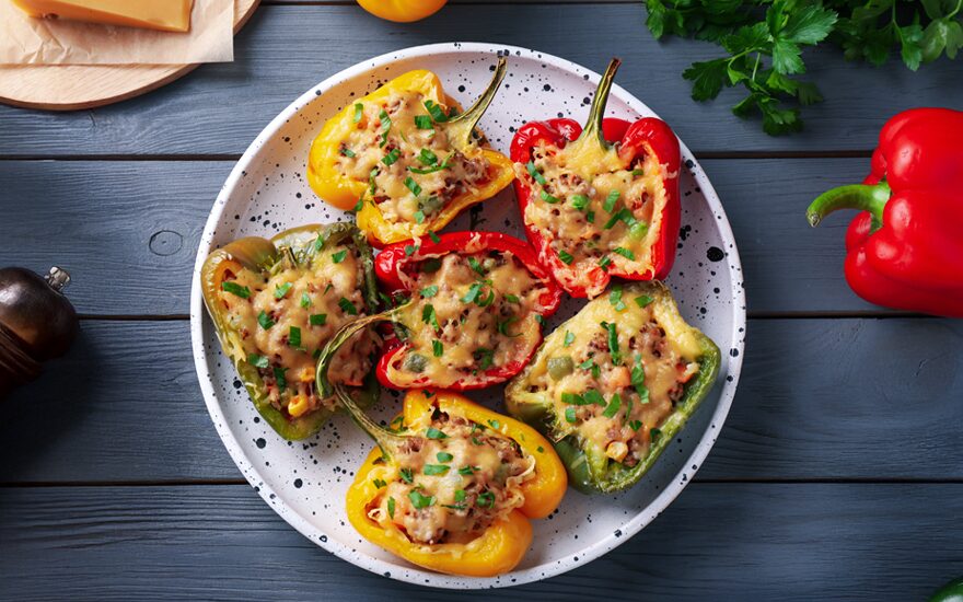 A plate of red, yellow, and green half stuffed peppers on a blue wooden table with ingredients around it.